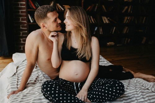 couple with one pregnant person in pajamas pants and a black sports bra sitting on their bed kissing and learning the best sex positions during pregnancy