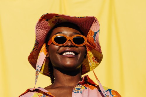 Portrait of happy young black female model in fashionable sunglasses and textile hat with floral print smiling brightly and looking away against yellow background