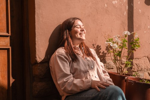 Portrait of a young redhead stylish woman wearing a pink jacket during a sunny day against a peach color clay wall in Cusco, Peru. She's resting while traveling South America.