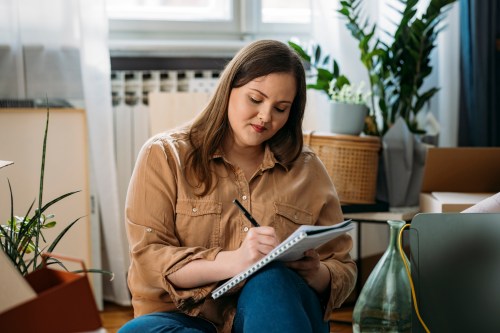 close up of person in tan long sleeve shirt and dark blue jeans sitting cross-legged on their living room floor writing in a pad of paper to learn about anxiety and procrastination