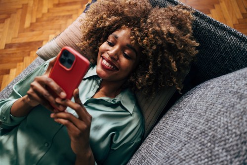 close up photo of a person with brown curly hair wearing a green long sleeve shirt lying down on their grey couch while looking at their phone for the Apollo Neuro vibration app sessions