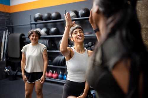 photo of two people high fiving and smiling at the best inclusive gyms, with a friends smiling in the background next to a stack of medicine balls and weights at the gym