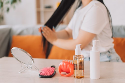 close up photo of haircare products, hairbrushes, and a mirror on a table while a person with long black hair applies gel to their hair in the background, hoping they don't use formaldehyde personal care products