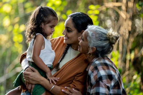 Three generations of women pose together outside for a portrait. A Mother holds her daughter in her arms as the Grandmother leans in closely and they share a laugh.