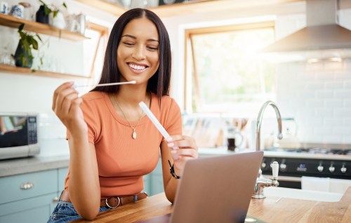 person wearing an orange short sleeve shirt and jeans holding a swab for the first home cervical cancer screening test that's been FDA approved while sitting at their kitchen table in front of their laptop