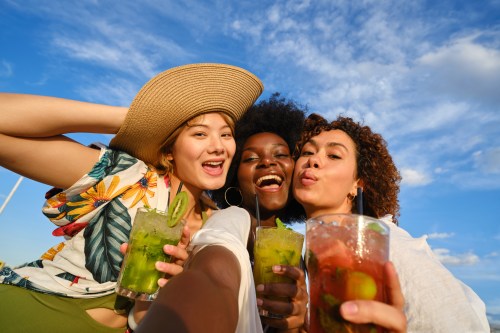 Frontal selfie view of three happy three women taking a selfie drinking cocktails outdoors in summer