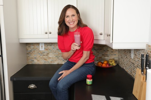 Molly Shannon sitting on a black marble countertop in a kitchen holding a pink smoothie.