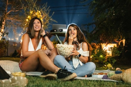 Nighttime photo of two people sitting outside eating popcorn using a projector to watch a summer rom-com
