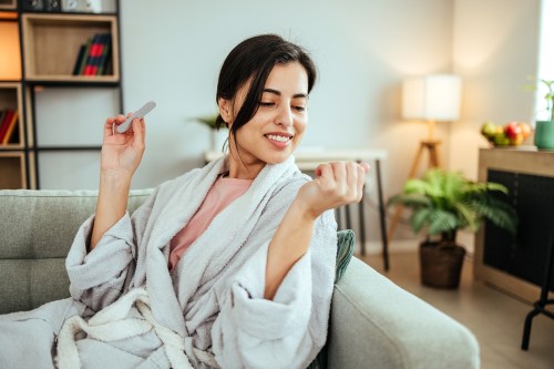 close of of person with dark brown hair in a low bun sitting on their couch in a light grey robe while filing their nails and looking for white spots on nails