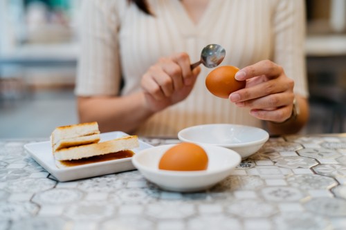 close up of person in a tan button up shirt sitting at the kitchen table cracking an egg with a spoon next to a plate of toast, wondering if they have an egg allergy