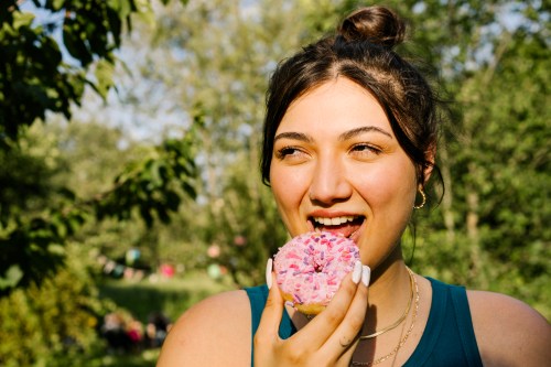 close up of person with brown hair up in a bun and a nose ring smiling while about to eat a bite of a donut with pink frosting and rainbow sprinkles, reaping the little treat benefits