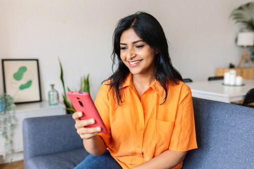 close up of person with long black hair in an orange short-sleeve button up shirt looking at their phone while sitting on their couch, hoping to use therapy by headspace app for affordable mental health care