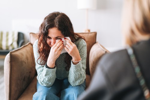close up of person with long brown hair wearing a mint green knit sweater and blue jeans sitting in a brown leather chair holding a tissue to their eyes while they cry, wondering why does your nose run when you cry